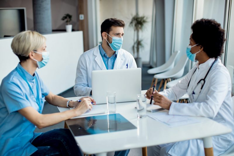 Doctor using laptop while communicating with female colleagues on a meeting at the clinic. They are wearing protective face masks due to coronavirus pandemic.